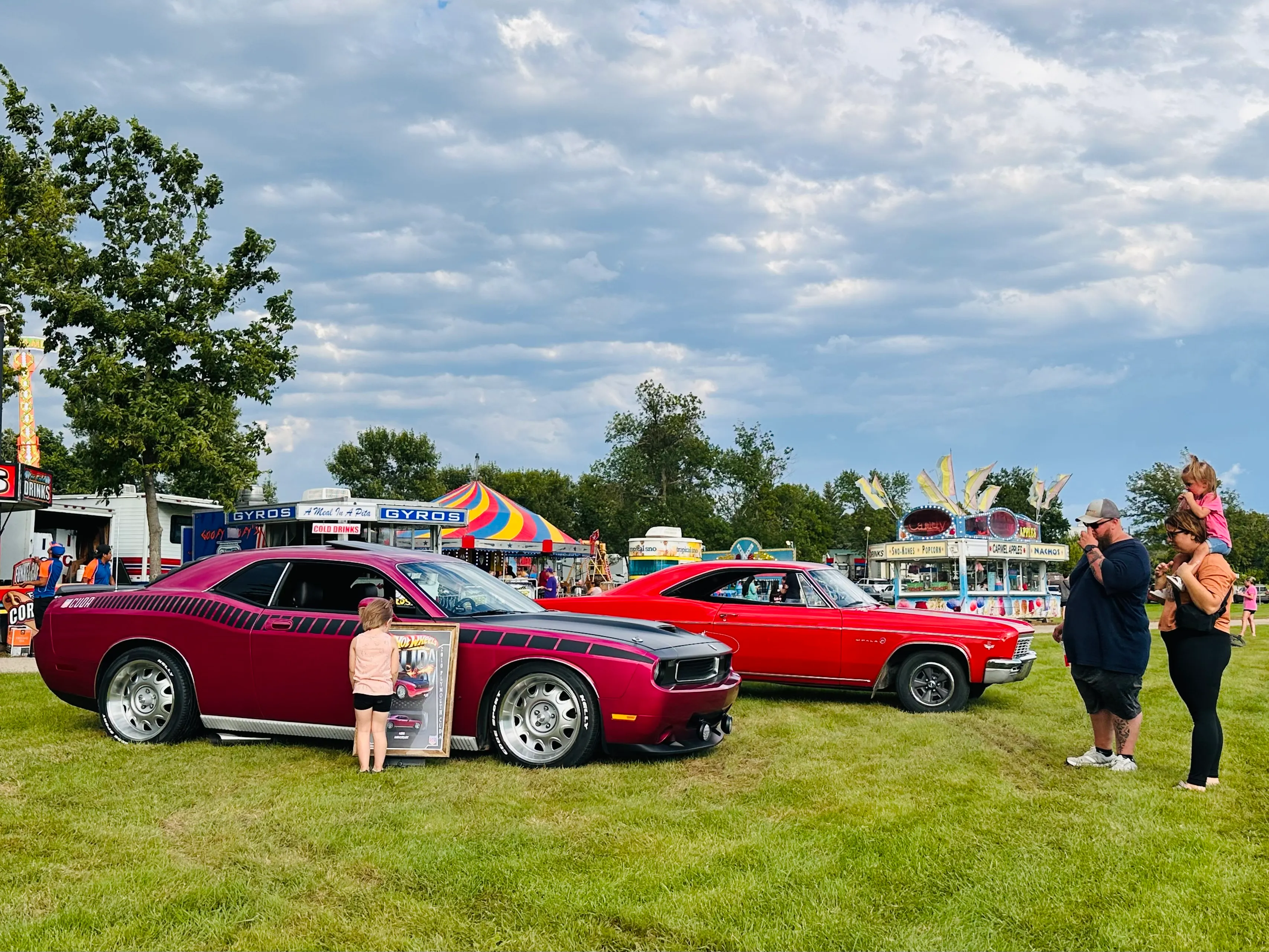 Classic red and maroon cars displayed on grass with carnival rides in background at the Wahpeton Round Up